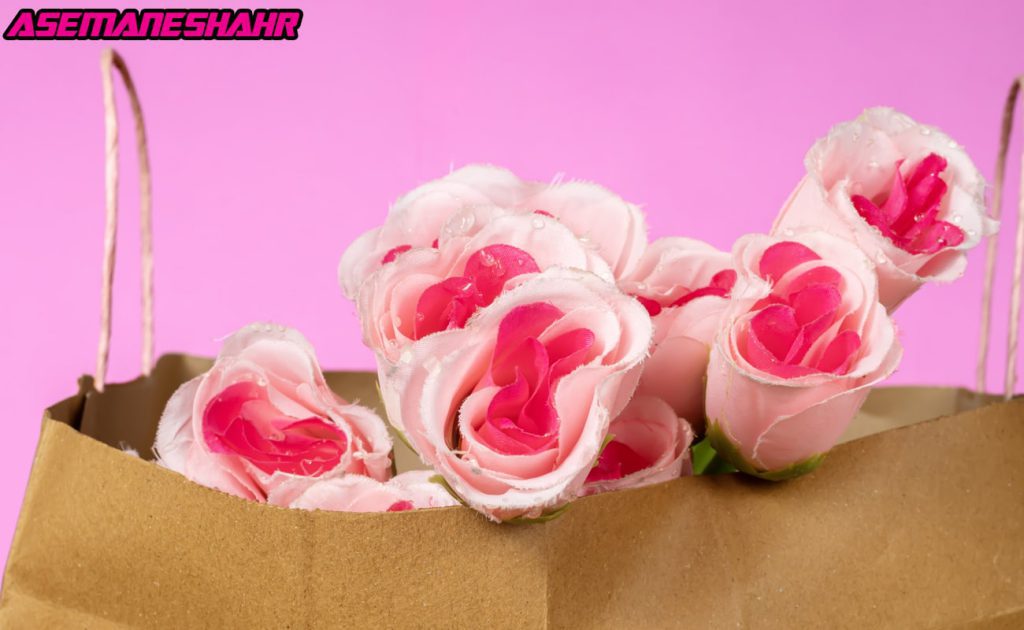 گل‌های رز صورتی مصنوعی با جزئیات پر زرق و برق، قرار گرفته داخل یک ساک دستی کاغذی، در پس‌ زمینه صورتی روشن، مناسب برای بسته‌ بندی هدایا عاشقانه.