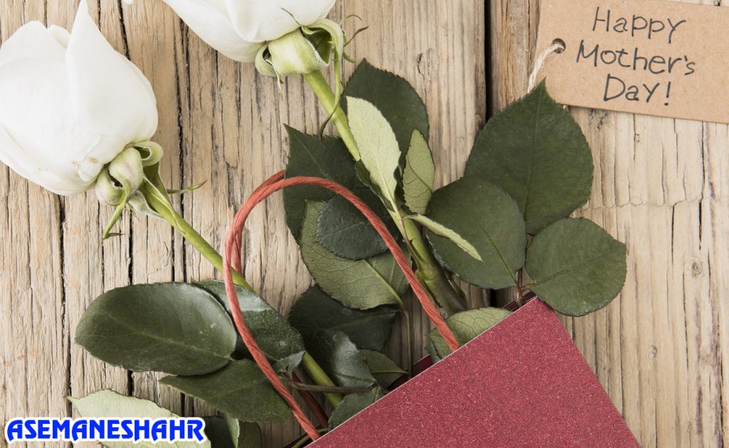 ساک دستی قرمز در کنار گل های رز سفید و کارت تبریک روز مادر | مناسب هدیه‌ ای خاص و احساسی برای مناسبت‌های ویژه.