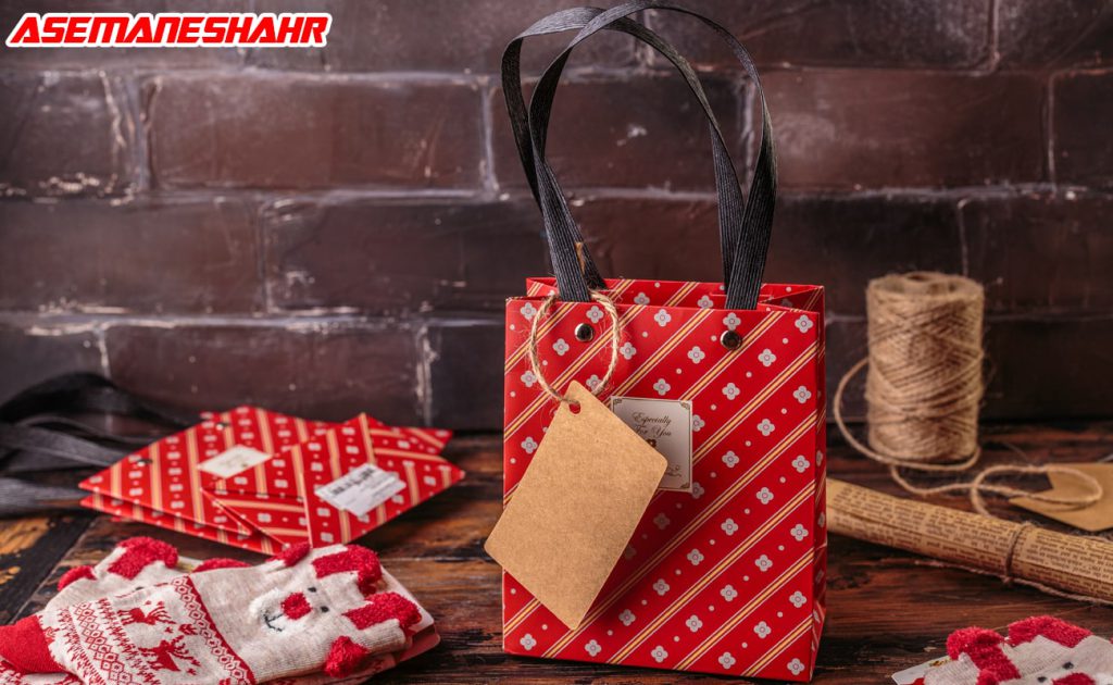 ساک کادویی با طراحی قرمز و سفید، حاوی برچسب کادویی، دستکش‌ های قرمز و سفید، و لوازم تزیینی دیگر در پس‌ زمینه چوبی تاریک.