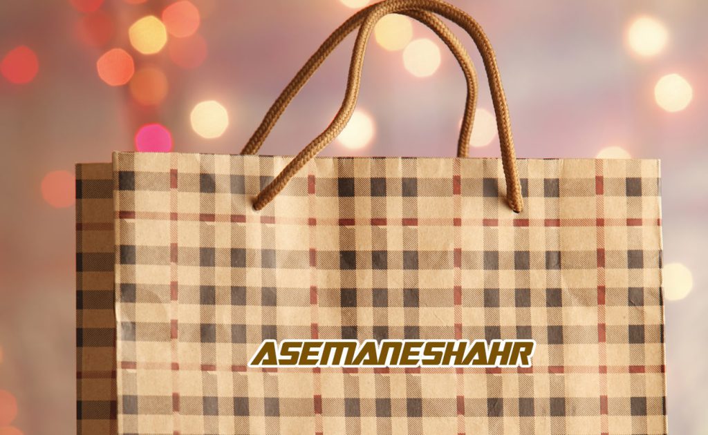 پاکت کادویی با طرح چهارخانه کلاسیک به رنگ کرم و قهوه‌ای، همراه با دسته‌های طنابی ضخیم و لوگوی آسمان شهر، مناسب برای بسته‌ بندی هدایا در مناسبت‌های مختلف.