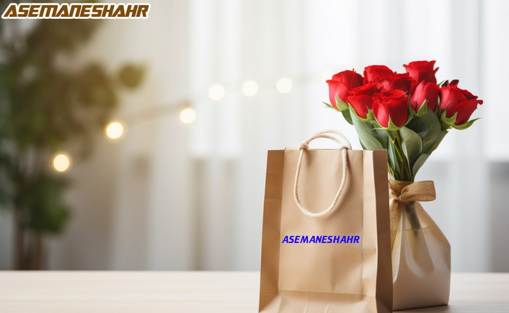 ساک کاغذی ساده با دسته کنفی و لوگوی Asemaneshahr، همراه با دسته‌ ای از گل‌های رز قرمز در گلدان برازنده، قرار گرفته روی میز چوبی با پس‌ زمینه‌ ای روشن و دکوراسیون لطیف.