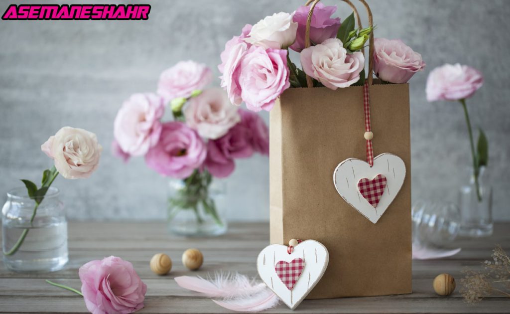 ساک دستی کاغذی با طراحی مینیمال تزئین‌ شده با قلب چوبی و گل‌های رز صورتی، قرار گرفته روی میز چوبی با گلدان‌های شیشه‌ ای.