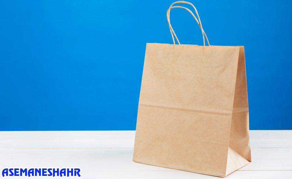 ساک دستی کرافت ساده با دسته کاغذی مقاوم روی میز سفید | مناسب برای بسته‌ بندی هدایا و خرید روزمره با پس‌ زمینه آبی روشن.