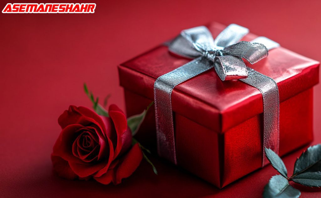 بسته هدیه قرمز با روبان نقره ای در کنار گل رز قرمز.