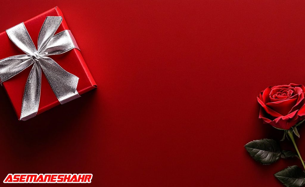 هدیه قرمز با روبان نقره ای در کنار رز قرمز زیبا.