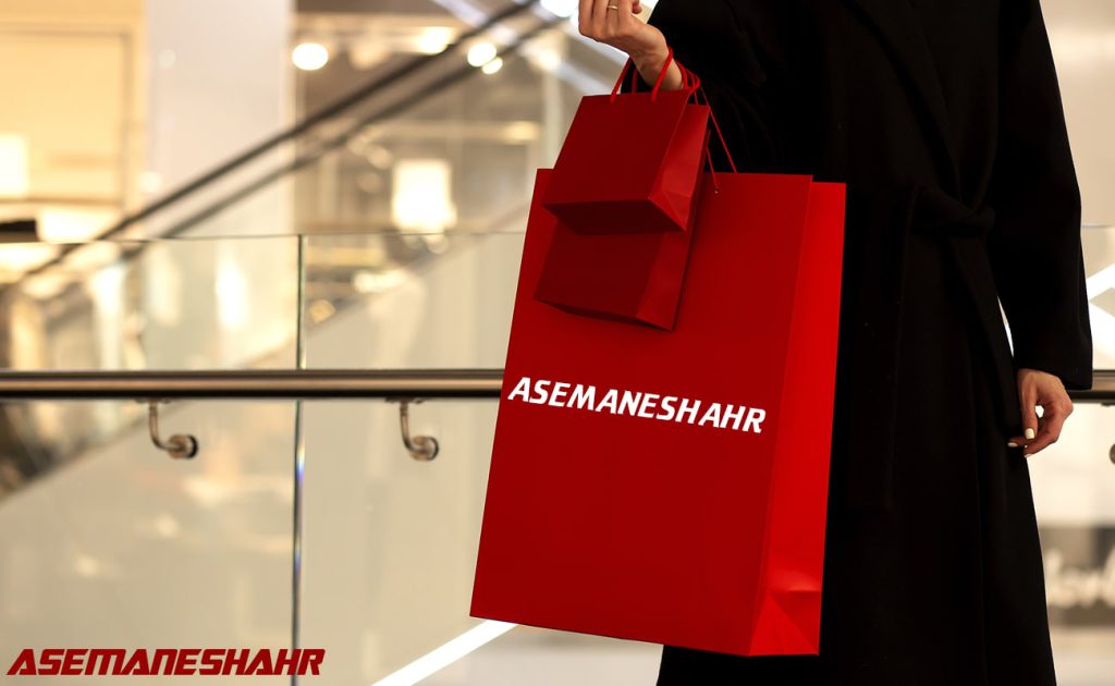 فردی با لباس مشکی در حال حمل دو ساک خرید قرمز با لوگوی Asemaneshahr، در محیط یک فروشگاه شیک و مدرن.