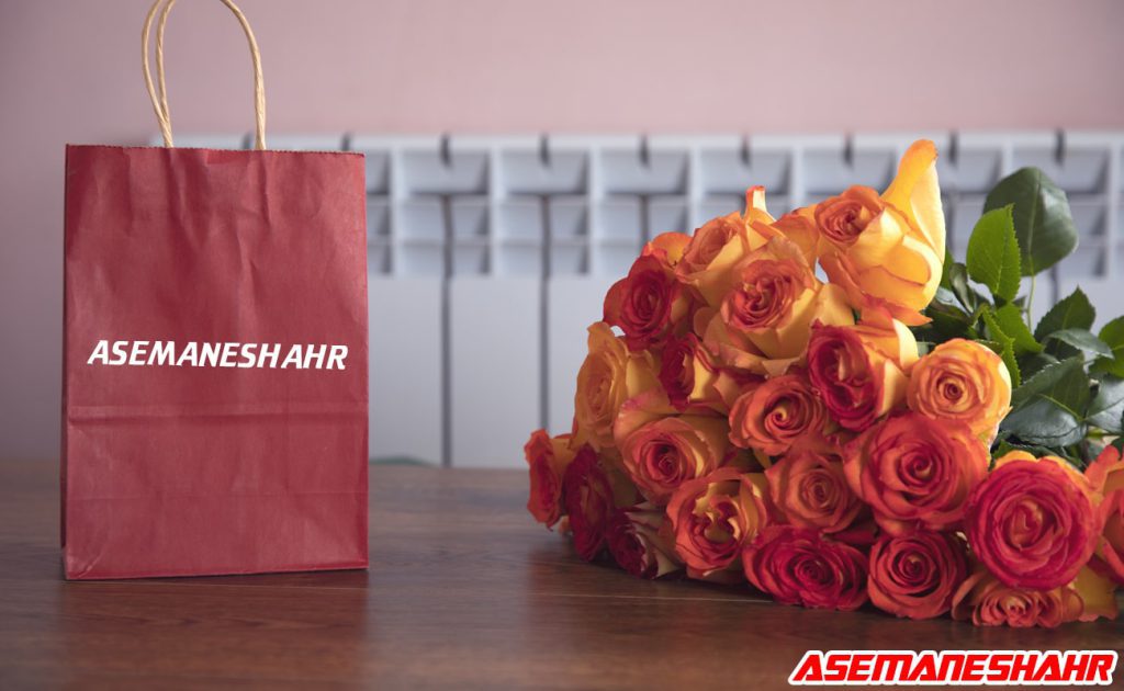 ساک دستی کاغذی قرمز با نام برند آسمان‌ شهر در کنار دسته گل رز نارنجی و قرمز، مناسب برای هدیه‌ های عاشقانه و مناسبت‌های ویژه.