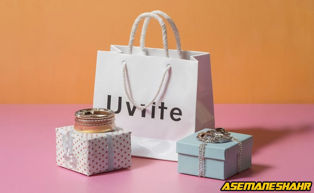 یک ساک دستی کاغذی سفید با نوشته “Uvite” به همراه یک جعبه کادویی خالدار با روبان نقره‌ای که چند حلقه روی آن قرار دارد و یک جعبه آبی روشن که یک گردنبند نقره‌ای روی آن است، بر روی یک سطح صورتی با پس‌ زمینه نارنجی قرار گرفته‌اند.