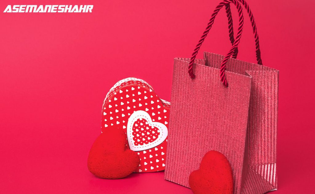  یک ساک دستی کاغذی بافت‌ دار قرمز، یک جعبه هدیه قلبی شکل با طرح قلب‌ های سفید، و دو قلب قرمز کوچک روی یک پس‌ زمینه قرمز.