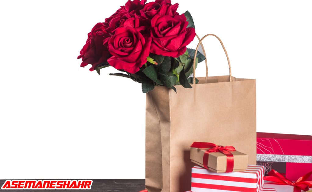 ساک دستی کاغذی با گل‌های سرخ در بالای آن و جعبه‌ های کادو با تزیینات قرمز و نقره‌ای در پس‌ زمینه سفید، مناسب برای هدیه‌ های عاشقانه.