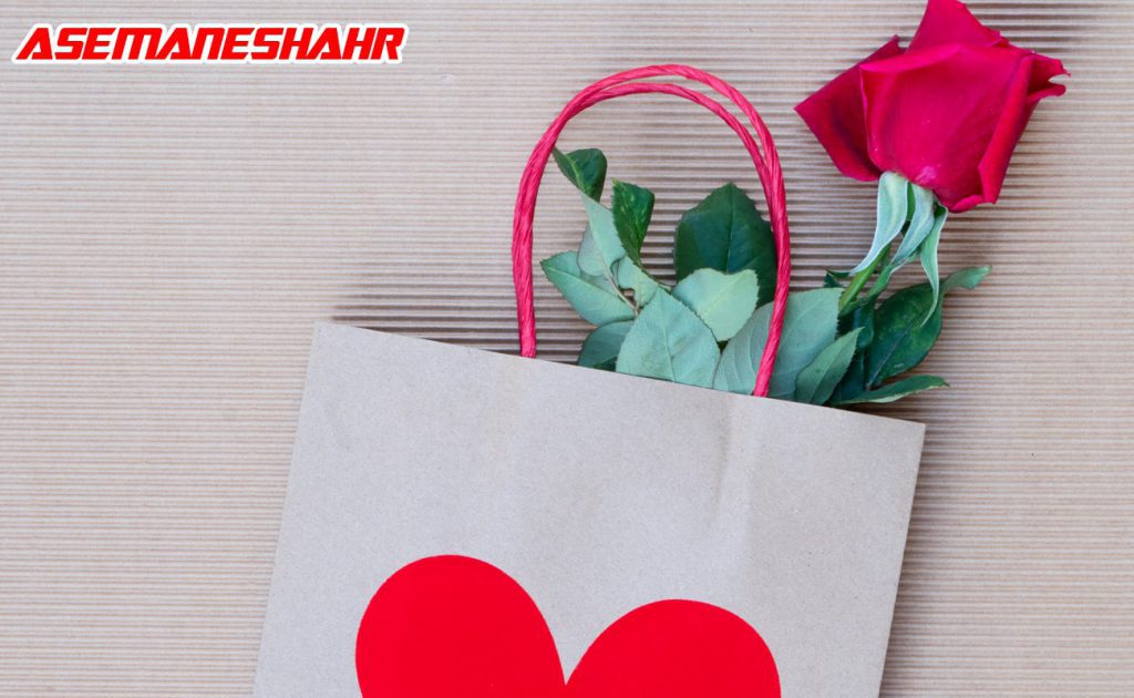 ساک دستی کاغذی با طرح قلب قرمز و دسته طنابی قرمز، همراه با گل رز قرمز که از داخل ساک بیرون آمده است.