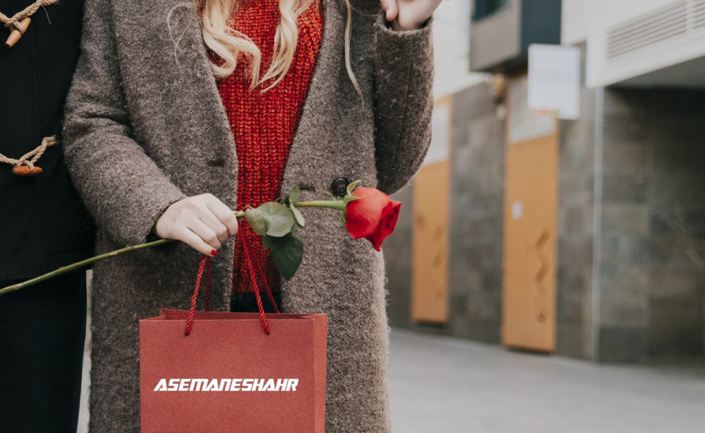خانمی با پالتو قهوه ای و باکس هدیه قرمز در دست، همراه با یک شاخه گل سرخ، مناسب برای هدیه دادن در عید نوروز.