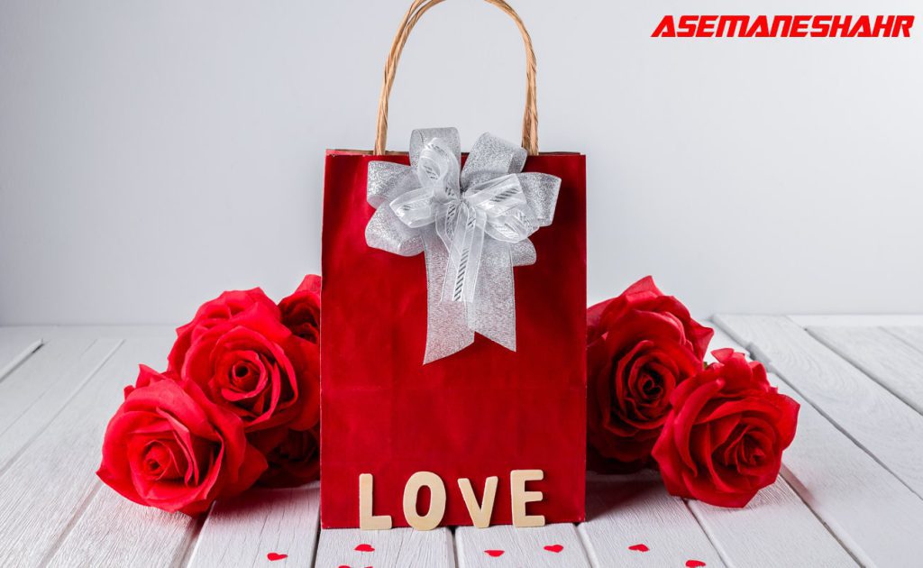 ساک دستی قرمز با روبان نقره‌ای، گل‌های رز قرمز، و حروف چوبی LOVE روی سطح چوبی سفید، مناسب برای هدیه ولنتاین یا مناسبت‌های عاشقانه.