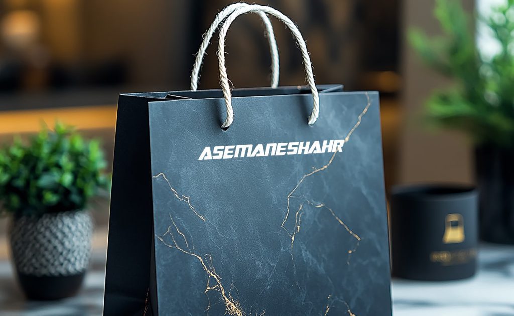 پاکت هدیه کاغذی شیک به رنگ مشکی با طرح مرمر و نوشته ASEMANSHAHR، همراه با دسته‌های طنابی، مناسب برای بسته‌بندی هدایای لوکس.