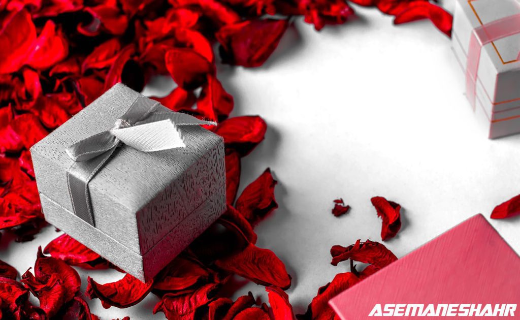 جعبه هدیه کوچک نقره‌ای همراه با روبان خاکستری در میان گلبرگ‌های خشک شده به رنگ قرمز، مناسب حال و هوای عاشقانه و روز ولنتاین.