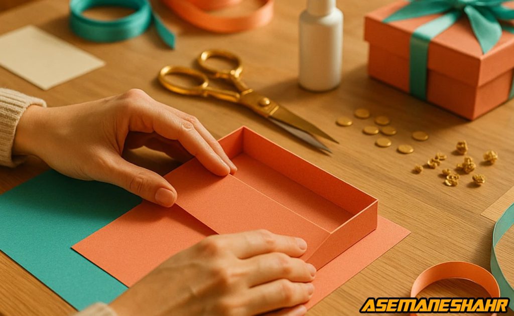 ساخت جعبه نارنجی و فیروزه‌ ای با دقت دست‌ ساز و ابزار طلایی روی میز کار چوبی در مراحل نهایی آموزش کادوپیچی.